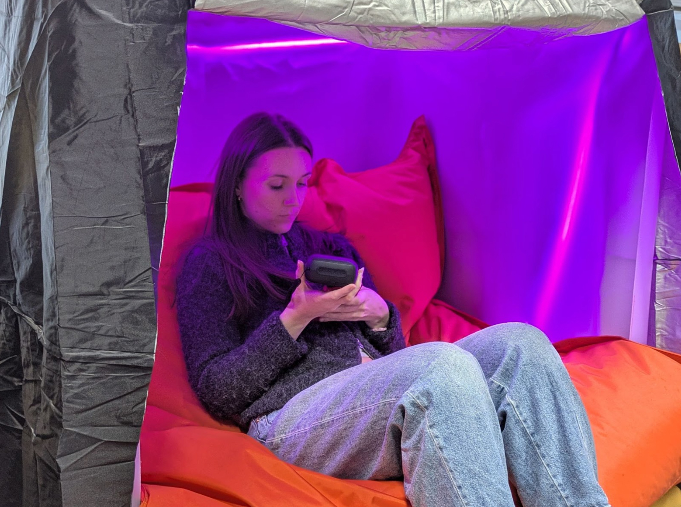 Une personne est assise à l'intérieur d'un cube sensoriel sur un très grand pouf orange qui maintient les jambes et le dos. La structure est composée d’une ancienne cuve de lait en parois en plastique blanc translucide, éclairée à l’intérieur par des lumières LEDs de couleur violette.  