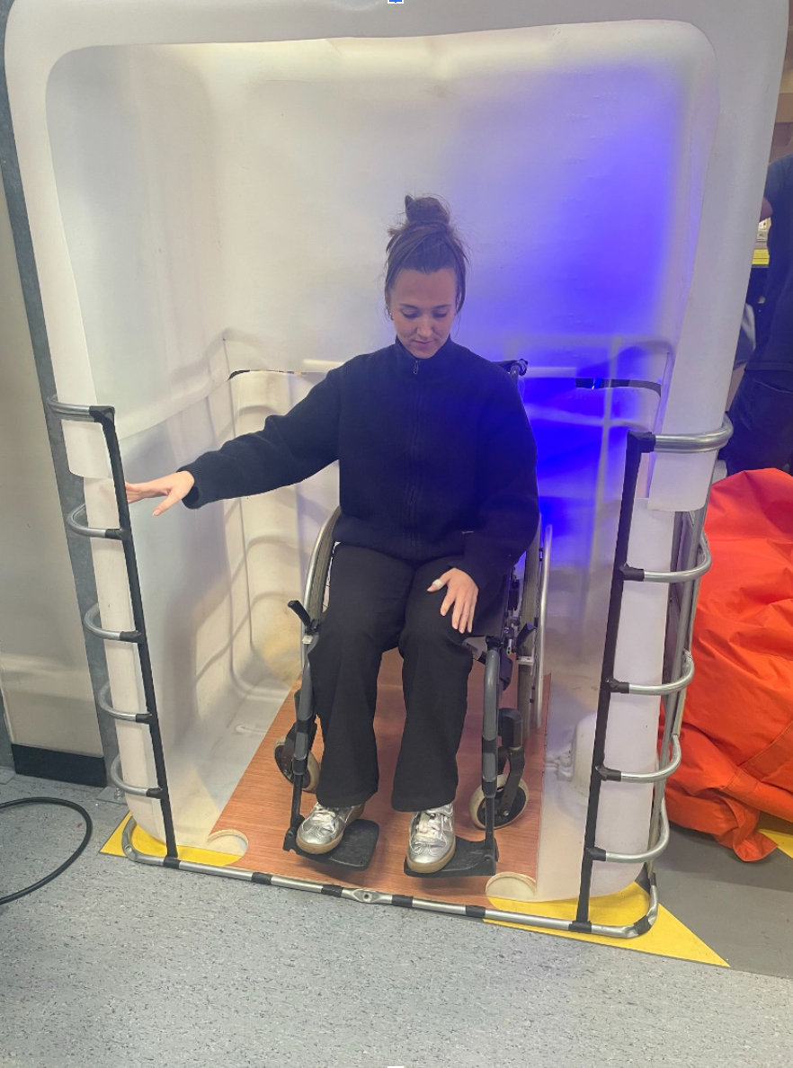 Une personne en fauteuil roulant à l'intérieur d'un cube sensoriel. La structure est composée d’une ancienne cuve de lait en parois en plastique blanc translucide.