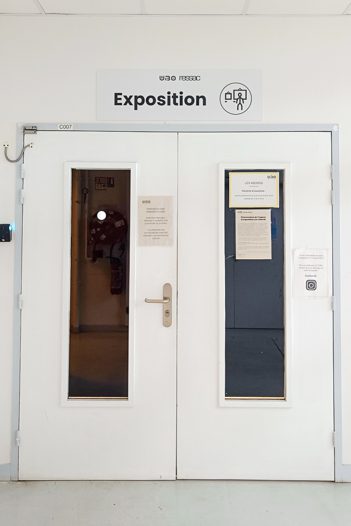 Entrée de l'espace Les Abords à la Faculté de Lettres et Sciences humaines de l'UBO avec au-dessus de la porte un panneau blanc avec un texte noir "Exposition" et un pictogramme noir symbolisant "Exposition"